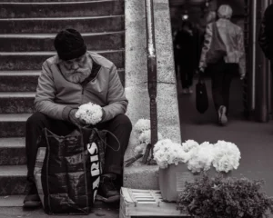 Cette photographie en noir et blanc met en scène un homme âgé assis sur une marche, le sujet principal. Il semble fatigué, les mains posées sur ses genoux.La composition est simple et centrée, avec un cadrage serré sur le vieil homme et ses fleurs blanches à côté de lui. En arrière-plan, une personne floue et un environnement urbain sont visibles.La lumière est douce, créant des contrastes modérés. Les tons dominants sont les gris et noirs, adoucis par les blancs des fleurs.Le style est documentaire et réaliste, évoquant une ambiance mélancolique et contemplative.