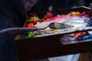 Cette photographie met en scène une palette de peintre couverte de peinture. Le sujet principal est la palette avec ses couleurs vives et mélangées, ainsi qu’un pinceau posé dessus.La composition est serrée, centrée sur la palette, avec un cadrage rapproché. La lumière semble naturelle, mettant en valeur les textures et les teintes. Les couleurs dominantes sont vives et variées : rouges, bleus, jaunes et verts.Le style évoque l’artistique et le créatif, avec une ambiance chaleureuse et expressive, typique d’un atelier de peinture.