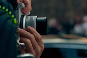 Cette photographie met en scène une personne tenant un appareil photo reflex numérique. Le sujet principal est les mains et l'appareil, avec un cadrage serré.La composition est simple et centrée sur le détail technique. La lumière semble naturelle, bien que légèrement tamisée, créant des contrastes doux. Les couleurs dominantes sont des tons neutres et froids, avec des nuances de gris et bleu.Le style est réaliste et documentaire, évoquant une ambiance professionnelle ou artistique. L'arrière-plan flou suggère un environnement public ou un événement, renforçant une impression de spontanéité et d'action.