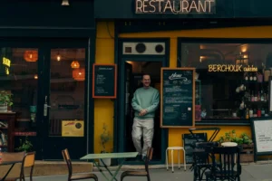Cette photographie met en scène un homme debout devant un restaurant nommé "Berghof". Le sujet principal est cet homme, vêtu d'un manteau sombre, positionné légèrement en contre-jour.La composition est symétrique avec des tables et des chaises disposées devant le restaurant. Le cadrage est serré, centré sur l'entrée éclairée. La lumière est douce et tamisée, créant une ambiance chaleureuse et nocturne. Les couleurs dominantes sont les tons jaunes et noirs du restaurant, contrastant avec les teintes sombres de la nuit.Le style est réaliste et urbain, évoquant une ambiance calme et intime.