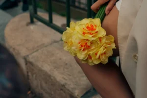 Cette photographie met en scène une personne tenant un bouquet de fleurs. Le sujet principal est le bouquet, composé de fleurs jaunes et orange, ainsi que le bras et la main de la personne, vêtue d’une robe blanche.La composition est simple et centrée, avec un cadrage serré sur le bouquet et le bras. La lumière est douce et naturelle, créant une ambiance chaleureuse et légèrement tamisée. Les couleurs dominantes sont les tons chauds des fleurs, contrastant avec le blanc de la robe.Le style est épuré et élégant, évoquant une ambiance romantique et intime.