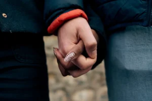 Cette photographie met en scène deux mains entrelacées, l’une portant une bague scintillante. Le sujet principal est l’alliance et le geste intime des mains.La composition est serrée, centrée sur les mains, avec un cadrage rapproché. La lumière est douce et naturelle, créant des ombres subtiles. Les couleurs dominantes sont les tons chauds (rouge du pull, bleu des jeans) et les teintes neutres.Le style est épuré et poétique, évoquant une ambiance tendre et romantique, presque intimiste.