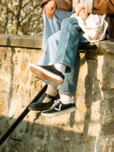 Cette photographie met en scène des **pieds et une partie de jambes** posés sur un muret en pierre. Le **sujet principal** est un individu vêtu de **jeans à carreaux** et de **sneakers noires**, avec un **pull blanc** en arrière-plan.Le **cadrage** est serré, centré sur les pieds et le bas des jambes, créant une composition minimaliste. La **lumière** est naturelle, douce et légèrement tamisée, suggérant un environnement extérieur en journée. Les **couleurs dominantes** sont les tons **bleus et blancs** des vêtements, contrastant avec le **gris-beige** du muret.L’ambiance est **détendue et décontractée**, évoquant une pause informelle en extérieur. Le style est **casual et moderne**.