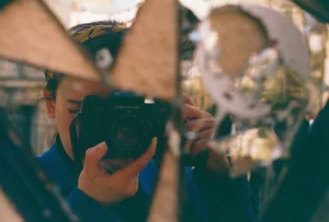 Cette photographie met en scène un photographe prenant une photo avec un appareil argentique. Le sujet principal est reflété dans un miroir brisé, créant une composition fragmentée et artistique.Le cadrage est serré, centré sur le visage et l’appareil. La lumière est douce et tamisée, donnant une ambiance intimiste et légèrement nostalgique. Les couleurs dominantes sont des tons chauds et neutres, avec des teintes bleutées et des reflets dorés.Le style évoque un mélange de réalisme et de surréalisme, renforcé par les reflets déformés. L’ambiance est à la fois contemplative et mystérieuse.