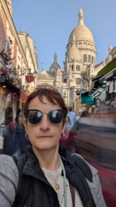Cette photographie montre une femme en selfie, sujet principal, portant des lunettes de soleil et une veste sombre. L’arrière-plan révèle une rue animée avec des bâtiments colorés et, au centre, la basilique du Sacré-Cœur de Paris, reconnaissable à son dôme blanc.Le cadrage est serré sur la personne, mais inclut une vue large de l’architecture. La lumière est naturelle et éclatante, typique d’un jour ensoleillé. Les couleurs dominantes sont les tons chauds des bâtiments et le blanc du Sacré-Cœur, créant une ambiance joyeuse et touristique. Le style est spontané et documentaire.