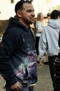 Cette photographie met en scène un homme comme sujet principal, vêtu d’un hoodie coloré avec un motif abstrait. Le cadrage est serré sur lui, avec des personnes floues en arrière-plan, suggérant un environnement urbain animé.La lumière semble naturelle, probablement en journée, avec des tons doux. Les couleurs dominantes sont les teintes vives du hoodie (bleu, violet, rose) contrastant avec les tons neutres des vêtements des autres personnes. Le style est spontané et réaliste, capturant un moment du quotidien. L’ambiance est décontractée et dynamique.