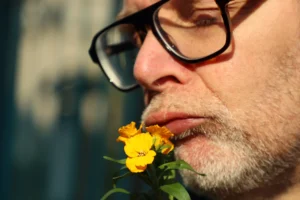 Cette photographie met en scène un homme comme sujet principal, qui semble humer une petite fleur jaune. Le cadrage est serré, centré sur son visage et la fleur, créant une intimité. La lumière naturelle, douce et légèrement contrastée, éclaire le sujet de face, mettant en valeur les détails.Les couleurs dominantes sont les tons chauds (jaune vif de la fleur, teints naturels de la peau) et les gris (vêtements, arrière-plan flou). Le style est simple et naturel, évoquant une ambiance poétique et contemplative, presque onirique.