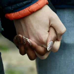 Cette photographie met en scène deux mains jointes, sujet principal. La composition est centrée et serrée, avec un cadrage rapproché qui isole les détails des doigts et des bracelets.La lumière est douce et naturelle, mettant en valeur les textures et les couleurs. Les teintes dominantes sont les tons chauds (orange du bracelet) et froids (peau, bleu des vêtements). Le style est épuré et réaliste, avec une ambiance intime et légèrement nostalgique, évoquant une connexion humaine subtile.