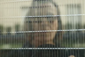 Cette photographie montre un visage flou vu à travers une structure métallique en treillis, probablement une cage ou une grille. Le sujet principal est indistinct, ce qui crée une impression de mystère.La composition est simple, centrée sur le visage partiellement caché. Le cadrage serré et les lignes horizontales de la grille dominent l'image, ajoutant une sensation de confinement.Les couleurs sont douces et neutres, avec des teintes grises et bleutées. La lumière semble naturelle mais tamisée, renforçant l'ambiance mélancolique et introspective. Le style est épuré et minimaliste, évoquant une atmosphère contemplative et légèrement mélancolique.