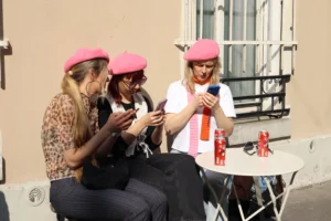 Cette photographie montre trois femmes assises, portant des bonnets roses et utilisant leurs téléphones. Le sujet principal est leur interaction avec les appareils, évoquant une ambiance moderne et détendue.La composition est symétrique, avec les femmes alignées autour d’une table. Le cadrage est serré, centré sur elles. La lumière est naturelle, probablement en extérieur, avec des ombres douces. Les couleurs dominantes sont les tons pastel (rose, beige) et les vêtements colorés.Le style est documentaire et spontané, capturant un moment du quotidien. L’ambiance est conviviale et décontractée.