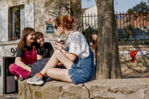 Cette photographie montre trois jeunes femmes assises sur un muret en pierre. Le sujet principal est leur interaction détendue, avec une ambiance conviviale et informelle.La composition est naturelle, avec un cadrage serré sur les trois personnes. La lumière est douce et naturelle, probablement en journée, éclairant les visages et créant des ombres légères. Les couleurs dominantes sont des tons vifs : rouge, bleu, vert et blanc.Le style est réaliste et spontané, évoquant une scène de vie quotidienne. L’ambiance est chaleureuse et décontractée, renforcée par les expressions et postures des jeunes femmes.