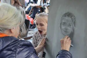 Cette photographie montre une scène de portrait en train d’être dessiné. Le sujet principal est une jeune fille observant son portrait en cours de réalisation sur une ardoise.La composition est serrée, mettant en valeur les interactions entre les personnes. Le cadrage est rapproché, centré sur les visages et les mains. La lumière semble naturelle, douce et légèrement tamisée. Les couleurs dominantes sont les tons froids (gris, bleu) et quelques touches chaudes (orange du vêtement).L’ambiance est conviviale et artistique, évoquant un moment de création et de partage.