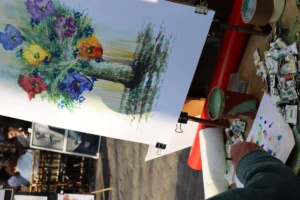 Cette photographie montre une personne tenant une grande toile colorée représentant un bouquet de fleurs stylisées et un paysage. Le sujet principal est cette peinture aux couleurs vives, dominées par des teintes bleues, violettes et vertes.La composition est dynamique, avec un cadrage serré sur la toile, mettant en valeur ses détails. La lumière semble naturelle, bien répartie, éclairant les couleurs sans ombre marquée. L’ambiance est artistique et créative, évoquant un marché ou une exposition d’art. Le style semble naïf ou artisanal, avec des traits expressifs et des couleurs saturées.