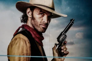 Cette photographie met en scène un homme incarnant un cow-boy, sujet principal. Il porte un chapeau, une veste en cuir et tient un revolver, évoquant un style western. La composition est centrée sur son visage et son arme, avec un cadrage serré. La lumière est naturelle et contrastée, mettant en valeur son expression déterminée. Les couleurs dominantes sont les tons chauds (beige, marron) et le rouge de sa veste, contrastant avec le ciel bleu clair. L’ambiance est dramatique et nostalgique, typique des films de western.