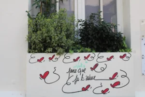 Cette photographie montre une fenêtre avec des graffitis en forme de cœurs et d'oiseaux rouges stylisés. Le sujet principal est l'inscription manuscrite en bas, partiellement illisible, avec des motifs décoratifs.La composition est simple et symétrique, centrée sur les motifs. Le cadrage est serré, mettant en valeur les détails des dessins. La lumière semble naturelle, éclairant uniformément la scène. Les couleurs dominantes sont les verts des plantes et les rouges des graffitis, créant un contraste vibrant.Le style est épuré et artistique, évoquant une ambiance à la fois poétique et un peu mélancolique.