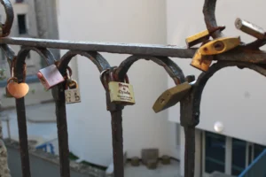 Cette photographie montre un ensemble de cadenas accrochés à une structure métallique, probablement une rambarde ou un garde-corps. Le sujet principal est ces cadenas, symboles souvent associés aux serments d’amour ou aux messages.La composition est serrée, avec un cadrage rapproché mettant en valeur les détails des cadenas. La lumière semble naturelle et douce, éclairant les objets sans ombres marquées. Les couleurs dominantes sont neutres (gris, métal) avec quelques touches de couleurs vives sur les cadenas (jaune, orange).Le style est documentaire et réaliste, créant une ambiance à la fois banale et poétique, évoquant la mémoire et les promesses.