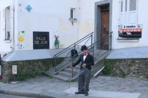 Cette photographie montre un homme en costume noir et chapeau, jouant de l'accordéon devant un immeuble. Le sujet principal est cet homme, situé au premier plan.La composition met en valeur les escaliers menant à une porte d’entrée, avec des enseignes ("Fille de joie" et "À vendre"). Le cadrage est serré sur l’artiste, avec un arrière-plan légèrement flou.La lumière est naturelle, douce, et les couleurs dominantes sont les tons neutres (gris, beige) avec des touches de vert et de bleu. L’ambiance est à la fois mélancolique et pittoresque, évoquant un quartier historique et artistique.