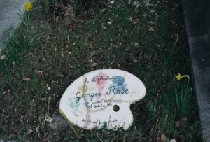 Cette photographie montre une palette de peintre en céramique personnalisée, posée sur une pelouse. Le sujet principal est la palette avec l'inscription "George's Rose" et une signature.La composition est simple et centrée, avec un cadrage serré. La lumière naturelle éclaire la scène, mettant en valeur les détails. Les couleurs dominantes sont les tons pastel de la palette (rose, bleu, vert) et le vert de la végétation. L’ambiance est calme et artistique, évoquant un hommage ou un souvenir lié à l’art.