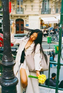 Cette photographie met en scène une femme comme sujet principal, assise sur une barrière en métal, vêtue d’une veste beige et d’une robe noire, tenant une fleur jaune. La composition est dynamique, avec un cadrage serré sur elle, mais incluant un arrière-plan urbain animé (bâtiments, vélos, passants).La lumière est naturelle et douce, typique d’une journée ensoleillée. Les couleurs dominantes sont les tons neutres (beige, gris) contrastés par des éclats de jaune et noir. Le style est à la fois naturel et élégant, évoquant une ambiance décontractée et chic en milieu urbain.