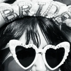 Cette photographie met en scène une personne portant un accessoire de fête avec le mot **« BRIDE »** en lettres scintillantes sur un bandeau et des lunettes en forme de cœur, également décorées.Le cadrage est serré sur le visage et les accessoires, créant une ambiance festive. La lumière est douce, mettant en valeur les détails brillants. Les couleurs dominantes sont le noir et blanc, avec des touches de blanc scintillant.Le style est rétro et ludique, évoquant une célébration joyeuse, probablement un mariage. L’ambiance est festive et romantique.