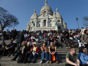 Cette photographie montre le **Sacré-Cœur de Montmartre** à Paris, avec sa basilique blanche et sa coupole bleue. Le **sujet principal** est la foule assise sur les marches, en plein air. La **composition** est dynamique, avec un premier plan occupé par les personnes et un arrière-plan monumental.Le **cadrage** est large, capturant à la fois les visiteurs et l’architecture. La **lumière** est naturelle et claire, suggérant un jour ensoleillé. Les **couleurs dominantes** sont les tons pastel de la basilique (blanc, bleu) et les vêtements colorés des gens.Le style est **documentaire et touristique**, évoquant une **ambiance conviviale et paisible**, typique d’un lieu emblématique et fréquenté.