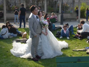 Cette photographie capture un couple, probablement des mariés, comme sujet principal. Le marié, en costume, et la mariée, en robe blanche à traîne, se tiennent au centre. Autour d’eux, des invités sont assis sur l’herbe, créant une ambiance conviviale et informelle.Le cadrage est large, incluant un espace vert avec des bâtiments en arrière-plan. La lumière est naturelle et ensoleillée, mettant en valeur les couleurs vives. Les tons dominants sont les blancs de la robe, les verts de la nature et les gris des vêtements. Le style est documentaire et spontané, évoquant une ambiance joyeuse et détendue.