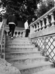 Cette photographie en noir et blanc capture un skateur descendant un escalier en pierre. Le sujet principal est le skateur en mouvement, au centre de l’image.La composition met en valeur les marches et la balustrade ornée. Le cadrage est serré sur les degrés, avec une perspective dynamique. La lumière naturelle, douce et contrastée, accentue les ombres et les textures. Les tons dominants sont les gris et noirs, renforçant l’ambiance urbaine et moderne.Le style est documentaire et spontané, évoquant une scène de vie quotidienne. L’ambiance est à la fois énergique et mélancolique, typique des clichés urbains en noir et blanc.