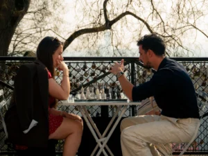 Cette photographie montre deux personnes assises à une table en métal, semblant discuter. La jeune femme, avec un sac à dos, est penchée légèrement en avant, tandis que l’homme, en chemise et pantalon clair, lui tend un objet.La composition est symétrique et centrée, avec un cadrage serré sur les deux sujets. La lumière naturelle, douce et légèrement dorée, évoque un moment en fin de journée. Les couleurs dominantes sont les tons neutres (beige, gris) et les touches de rouge et bleu. Le style est naturel et spontané, créant une ambiance intime et chaleureuse.
