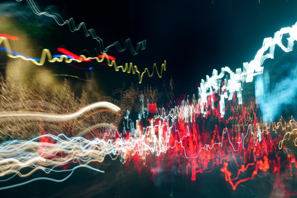 Cette photographie capture des **traînées lumineuses** en mouvement, probablement des phares et feux de véhicules. Le **sujet principal** est l’énergie dynamique des lignes colorées, créant un effet de vitesse.La **composition** est fluide, avec des courbes et des trajectoires variées. Le **cadrage** est large, englobant une scène nocturne urbaine.Les **couleurs dominantes** sont des **tons chauds** (rouge, orange) et des **teintes froides** (bleu, blanc), formant un contraste vibrant. Le **style** relève de la photographie longue exposition, évoquant **mouvement et énergie**.L’**ambiance** est **rythmée et électrique**, transmettant une sensation de vitesse et de vie nocturne intense.