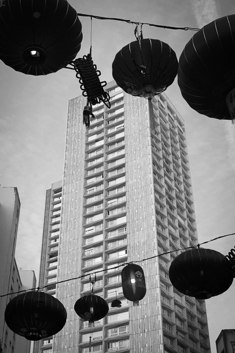 Thierry · 27/01/2024 · 2560 x 3840 Cette photographie en noir et blanc met en scène un immeuble moderne en hauteur, sujet principal. La composition joue sur la perspective verticale, accentuée par des pots de fleurs suspendus en premier plan, créant une répétition graphique.Le cadrage est serré vers le bas, avec une lumière naturelle diffuse, probablement en journée. Les ombres sont marquées, renforçant le contraste. Les couleurs dominantes sont les nuances de gris, typiques du noir et blanc, avec des touches de blanc et de noir pur.Le style est artistique et urbain, évoquant une ambiance à la fois moderne et nostalgique.