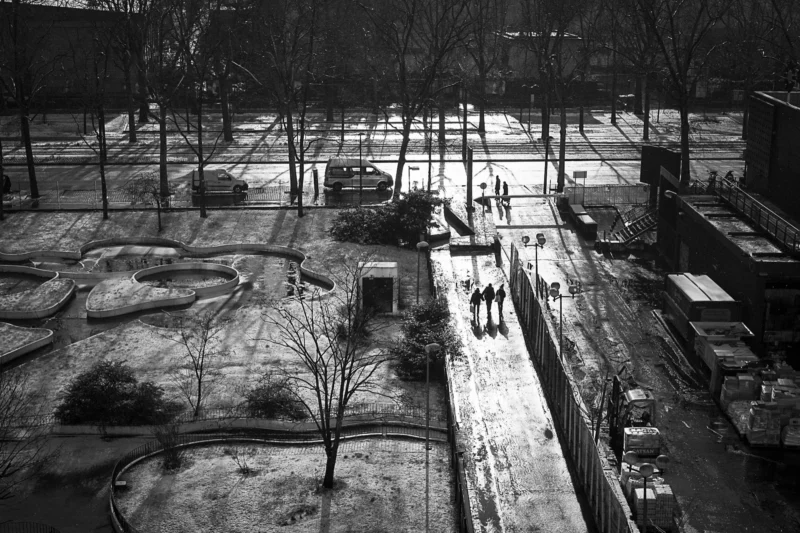 Thierry · 18/01/2024 · 3840 x 2560 Cette photographie en noir et blanc capture un parc enneigé vu d'en haut. Le sujet principal semble être l'espace urbain et ses usagers, avec quelques personnes marchant sur un chemin. La composition est aérée, avec des lignes droites et des formes géométriques (allées, bâtiments). Le cadrage large englobe une vue aérienne, offrant une perspective panoramique.La lumière, bien que diffuse, accentue les contrastes entre ombres et zones éclairées. Les couleurs dominantes sont les nuances de gris, renforçant l'atmosphère hivernale. Le style est documentaire et épuré, évoquant une ambiance calme et solitaire, presque mélancolique.