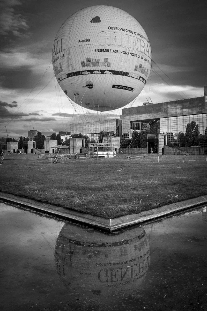 Thierry · 01/01/2018 · 2555 x 3840 Cette photographie met en scène un ballon dirigeable suspendu au-dessus d’une ville. Le sujet principal est ce ballon, dont les détails et publicités sont visibles. La composition joue sur la perspective avec un cadrage bas, créant un effet de distorsion et de grandeur. La lumière, naturelle et contrastée, accentue les ombres et les textures, renforçant le côté dramatique. En noir et blanc, les tons dominants varient du blanc éclatant au noir profond, soulignant les contrastes urbains. Le style est artistique et cinématographique, évoquant une ambiance mystérieuse et futuriste.