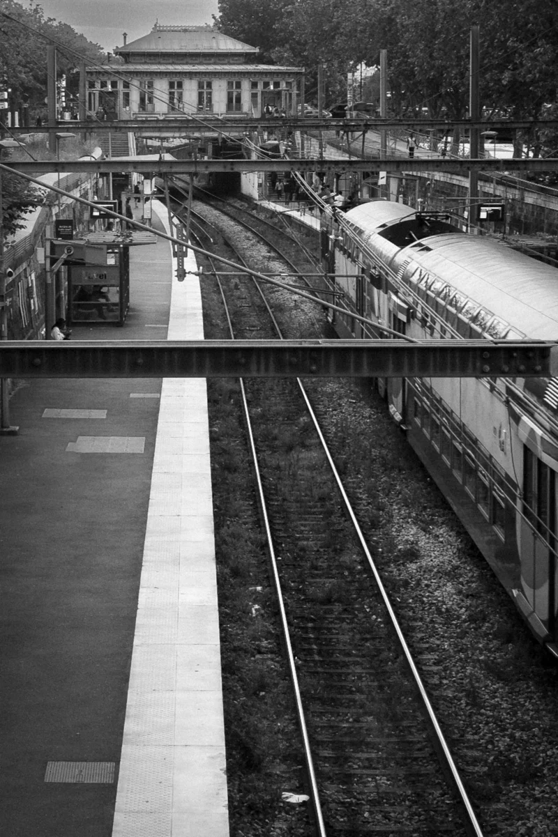 Thierry · 01/01/2018 · 2559 x 3840 Cette photographie en noir et blanc capture une gare ferroviaire. Le sujet principal est un train stationné sur une voie, avec une plateforme déserte en premier plan. La composition met en valeur les rails convergeant vers l’horizon, encadrés par des bâtiments traditionnels en arrière-plan.Le cadrage est légèrement en contre-plongée, accentuant la perspective. La lumière, douce et diffuse, crée des contrastes marqués entre ombres et clartés. L’ambiance est à la fois calme et nostalgique, renforcée par l’absence de couleurs et de mouvement. Le style rappelle le photojournalisme classique.