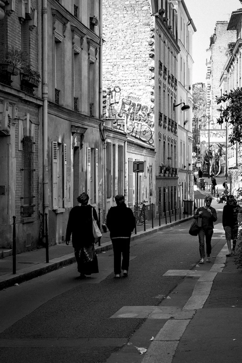 Thierry · 14/04/2024 · 2560 x 3840 Cette photographie en noir et blanc capture une rue étroite et urbaine. Le sujet principal est un groupe de trois personnes marchant, dont une pousse un landau. La composition met en valeur les bâtiments anciens et les détails architecturaux, avec une perspective convergente vers l'arrière-plan.Le cadrage serré accentue l'intimité de la scène. La lumière, douce et naturelle, crée des contrastes marqués entre ombres et clartés, renforçant l'atmosphère. Les tons dominants sont les gris et noirs, typiques du monochrome, ajoutant une touche nostalgique et dramatique. Le style est documentaire et réaliste, évoquant une ambiance calme et quotidienne dans un cadre historique.