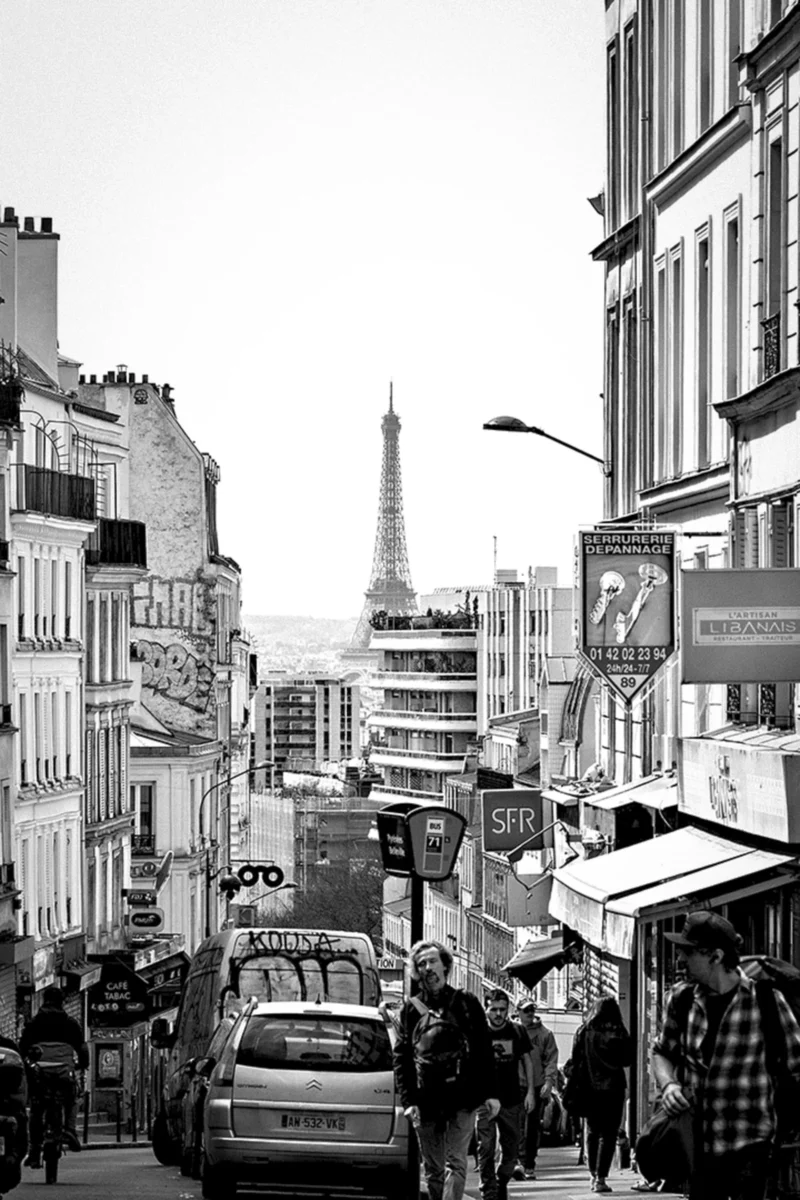Thierry · 14/04/2024 · 2560 x 3840 Cette photographie en noir et blanc capture une rue parisienne animée. Le **sujet principal** est l’**avenue menant à la Tour Eiffel**, visible au loin. La **composition** est dynamique, avec une perspective convergente vers l’horizon.Le **cadrage** est serré, mettant en valeur les bâtiments, les passants et les voitures. La **lumière** est naturelle, douce, créant des contrastes nets. Les **couleurs dominantes** sont les gris et noirs, typiques du noir et blanc, accentuant les textures et ombres.Le **style** est documentaire et urbain, évoquant une **ambiance** vivante et typiquement parisienne, mêlant quotidien et monumentalité.