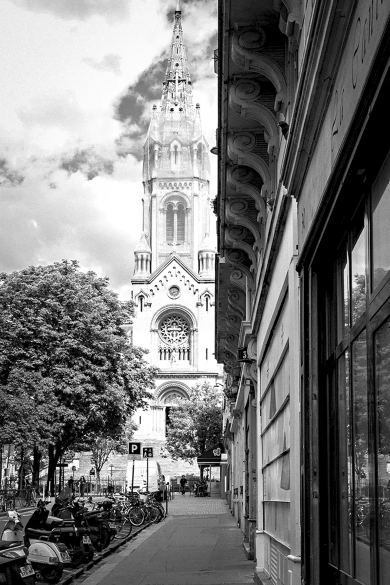 Thierry · 26/05/2024 · 2560 x 3840 Cette photographie met en scène une **église gothique** comme sujet principal, avec sa façade imposante et ses vitraux. La **composition** joue sur la perspective, encadrée par un bâtiment moderne à droite, créant une ligne verticale dynamique.Le **cadrage** est serré, guidant le regard vers le ciel. La **lumière**, douce et naturelle, souligne les contrastes entre ombres et clartés, typique d’un jour nuageux. En **niveau de couleurs**, le rendu est en **noir et blanc**, accentuant les textures et les jeux d’ombres.Le style est **classique et intemporel**, évoquant une ambiance **sereine et mélancolique**, mêlant histoire et modernité urbaine.