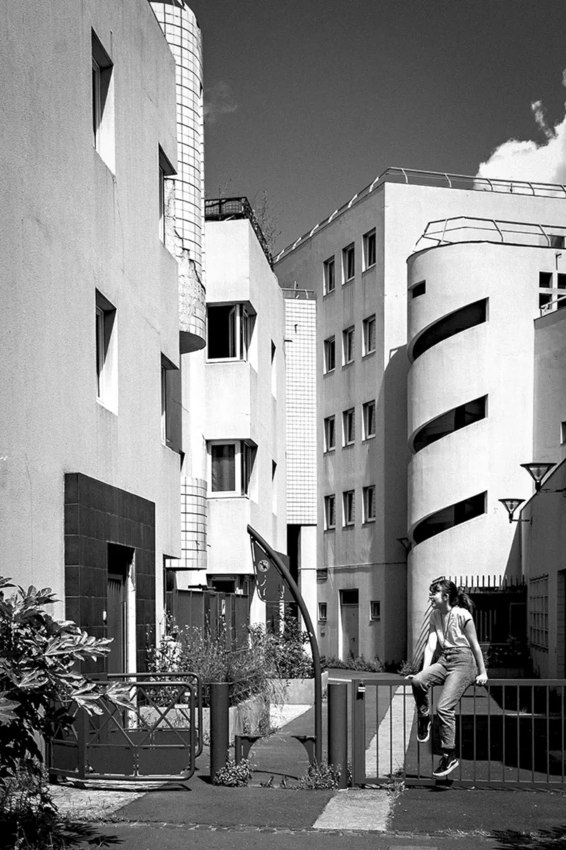 Thierry · 26/05/2024 · 2560 x 3840 Cette photographie en noir et blanc met en scène un homme assis sur une rampe d’escalier extérieure, devenant le sujet principal. La composition joue sur les lignes architecturales géométriques des bâtiments modernes aux formes anguleuses et courbes. Le cadrage est serré, centré sur l’individu et son environnement immédiat.La lumière, naturelle et contrastée, accentue les ombres et les volumes. Les couleurs dominantes, bien que monochromes, varient du blanc au gris foncé. Le style architectural semble contemporain et minimaliste, créant une ambiance à la fois urbaine et introspective.