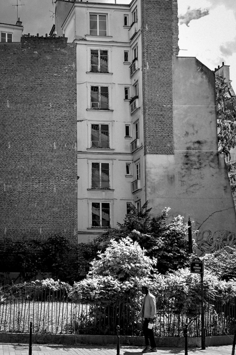 Thierry · 26/05/2024 · 2560 x 3840 Cette photographie en noir et blanc met en scène un immeuble parisien typique, sujet principal, avec ses fenêtres alignées et sa façade légèrement dégradée. La composition joue sur le contraste entre l’architecture urbaine et la nature environnante, avec un arbre et un petit espace vert en premier plan.Le cadrage est serré sur l’immeuble, encadré par la végétation. La lumière, douce et diffuse, adoucit les contours, créant une ambiance nostalgique et mélancolique. Les tons gris dominent, accentuant le côté intemporel et poétique de la scène, typique du style photographique documentaire ou artistique des époques passées.