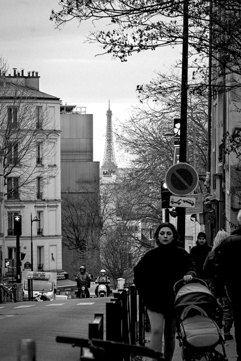 Thierry · 27/12/2023 · 2560 x 3840 Cette photographie en noir et blanc capture une rue parisienne animée. Le sujet principal est la Tour Eiffel, visible au loin, encadrée par des immeubles haussmanniens. La composition joue sur la perspective avec une ligne de fuite vers l’horizon.Le cadrage inclut des passants, des arbres et des éléments urbains comme des feux tricolores. La lumière est douce, typique d’un jour couvert, adoucissant les contrastes. Les couleurs dominantes, bien que monochromes, varient des gris clairs aux noirs profonds.Le style est documentaire et réaliste, évoquant une ambiance urbaine paisible et mélancolique.