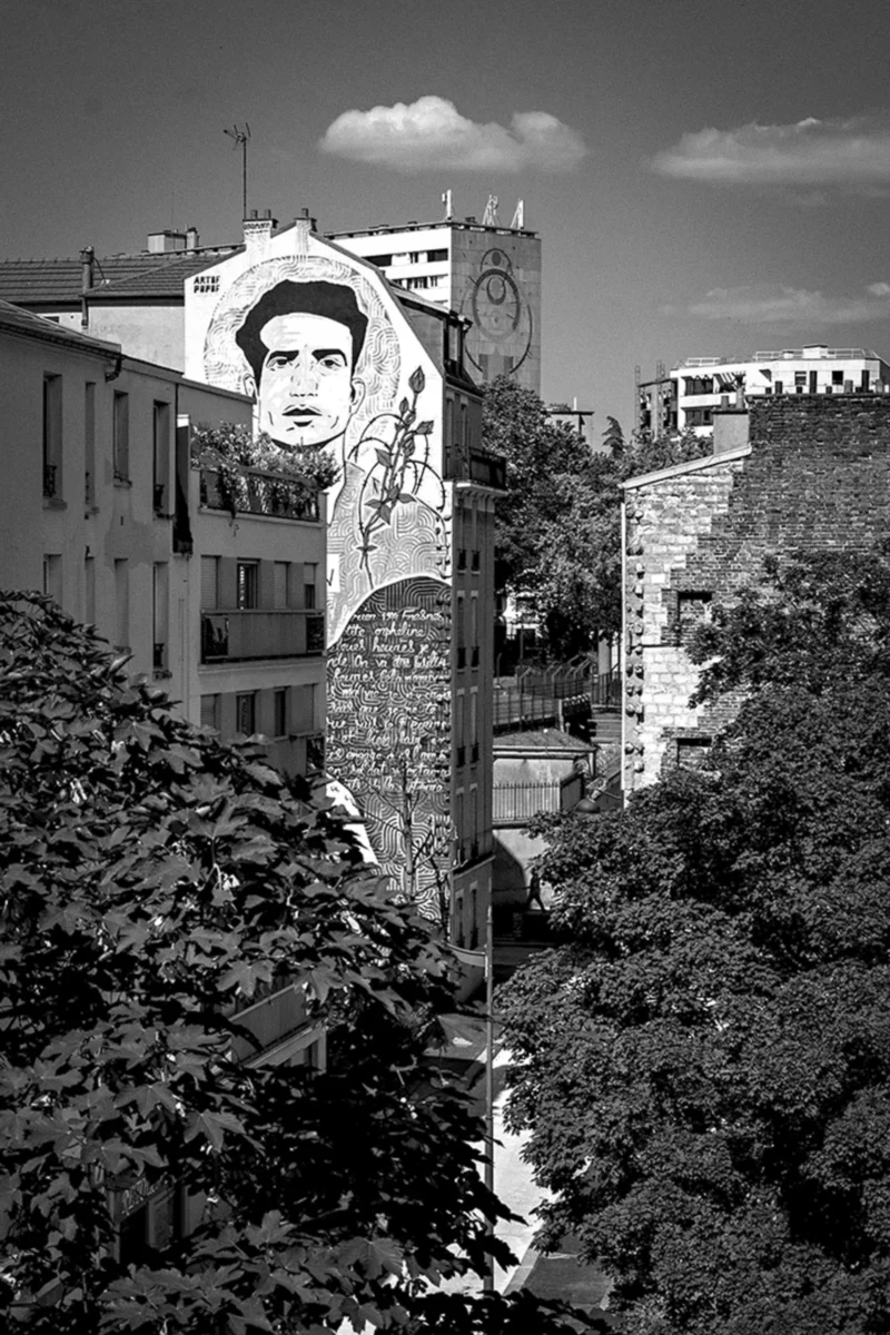 Thierry · 09/06/2024 · 2560 x 3840 Cette photographie en noir et blanc met en scène un **mural géant** représentant un visage masculin, probablement une figure historique ou politique. La **composition** est verticale, avec un **cadrage serré** sur le mural, partiellement caché par des **feuillages en premier plan**.La **lumière** est naturelle, contrastée, accentuant les textures des bâtiments et des arbres. Les **couleurs dominantes** sont les nuances de gris, renforçant l’aspect dramatique et intemporel de l’image. Le **style** est documentaire, avec une touche artistique due au choix du sujet et du cadrage. L’**ambiance** est à la fois mystérieuse et solennelle, évoquant une réflexion sur la mémoire et l’Histoire urbaine.