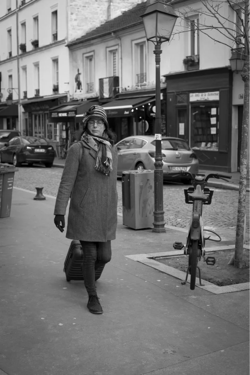 Thierry · 04/07/2025 · 2560 x 3840 Cette photographie en noir et blanc met en scène une femme comme sujet principal, vêtue d’un long manteau et d’un chapeau, marchant sur un trottoir urbain. La composition est simple et équilibrée, avec un cadrage serré sur elle, tout en intégrant des éléments architecturaux et urbains en arrière-plan.La lumière est douce, probablement naturelle, créant des contrastes modérés. Les tons dominants sont les gris et noirs, typiques du noir et blanc, accentuant les textures et les ombres. Le style évoque une ambiance rétro, peut-être des années 1950-1960, avec une touche mélancolique et intemporelle.