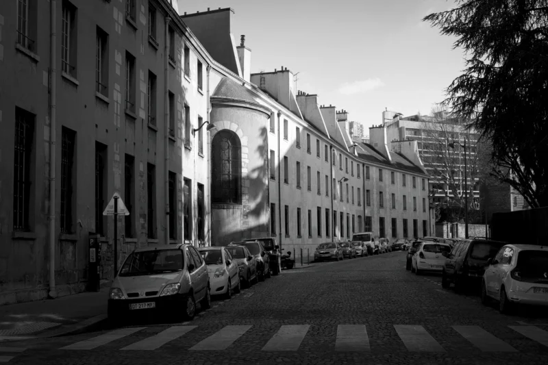 Thierry · 04/07/2025 · 3840 x 2560 Cette photographie en noir et blanc capture une rue urbaine étroite et calme. Le sujet principal est l’architecture des bâtiments anciens, alignés de part et d’autre de la voie. La composition met en valeur une perspective convergente vers l’horizon, renforcée par les voitures garées et les arbres en fond.Le cadrage est serré, centré sur les façades et les détails architecturaux. La lumière, douce et diffuse, crée des contrastes modérés, accentuant les textures des murs et des véhicules. Les tons dominants sont les gris et noirs, typiques du monochrome, évoquant une ambiance à la fois nostalgique et intemporelle, avec une touche mélancolique.