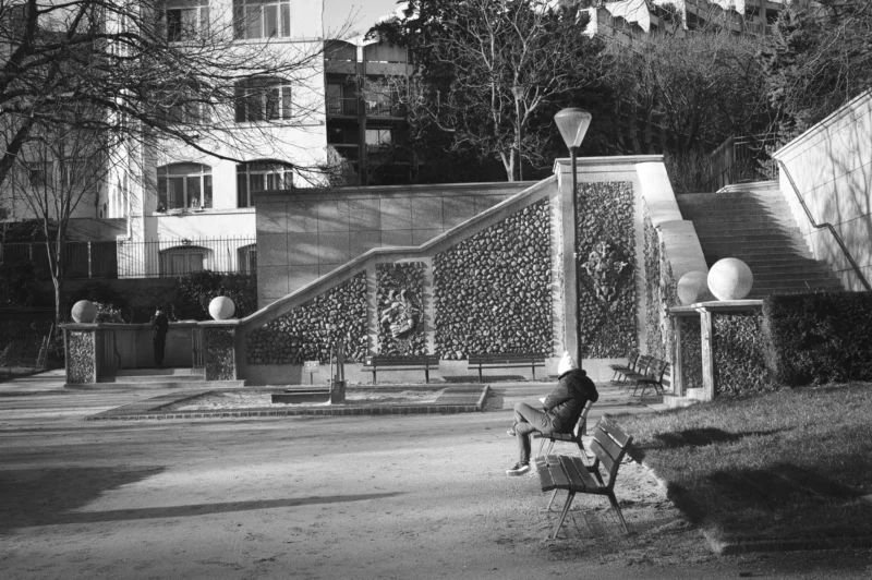 Thierry · 04/07/2025 · 3840 x 2555 Cette photographie en noir et blanc met en scène un **sujet principal** : un homme assis sur un banc, seul, dans un parc.La **composition** joue sur les lignes des escaliers et des allées, créant une perspective profonde. Le **cadrage** inclut des éléments architecturaux et naturels, comme les bâtiments en arrière-plan et les arbres.La **lumière**, douce et tamisée, accentue les contrastes et donne une ambiance mélancolique. Les **couleurs dominantes** sont les nuances de gris, renforçant l’aspect intemporel.Le **style** est documentaire et contemplatif, évoquant une **ambiance** calme et solitaire, presque nostalgique.