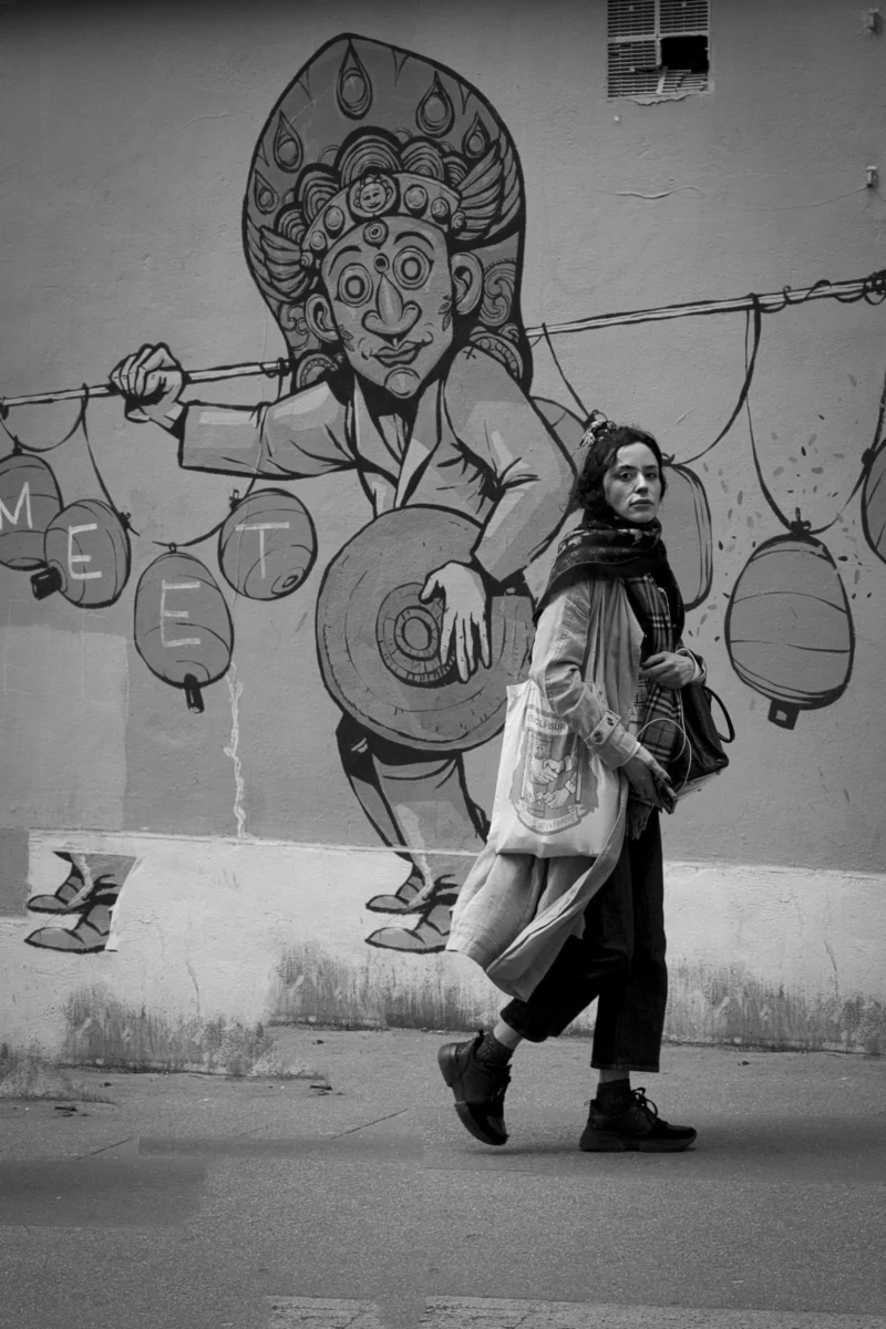 Thierry · 04/07/2025 · 2560 x 3840 Cette photographie en noir et blanc met en scène deux personnes marchant devant un mur tagué. Le sujet principal est le graffiti mural représentant une figure stylisée tenant des objets suspendus, dont des ballons et des instruments de musique.La composition joue sur le contraste entre les passants et l’art urbain. Le cadrage est serré, capturant les détails du graffiti et les silhouettes en mouvement. La lumière est naturelle, créant des ombres marquées et un contraste fort. L’ambiance est à la fois urbaine et artistique, avec une touche de mélancolie due au monochrome. Le style est street art, dynamique et expressif.