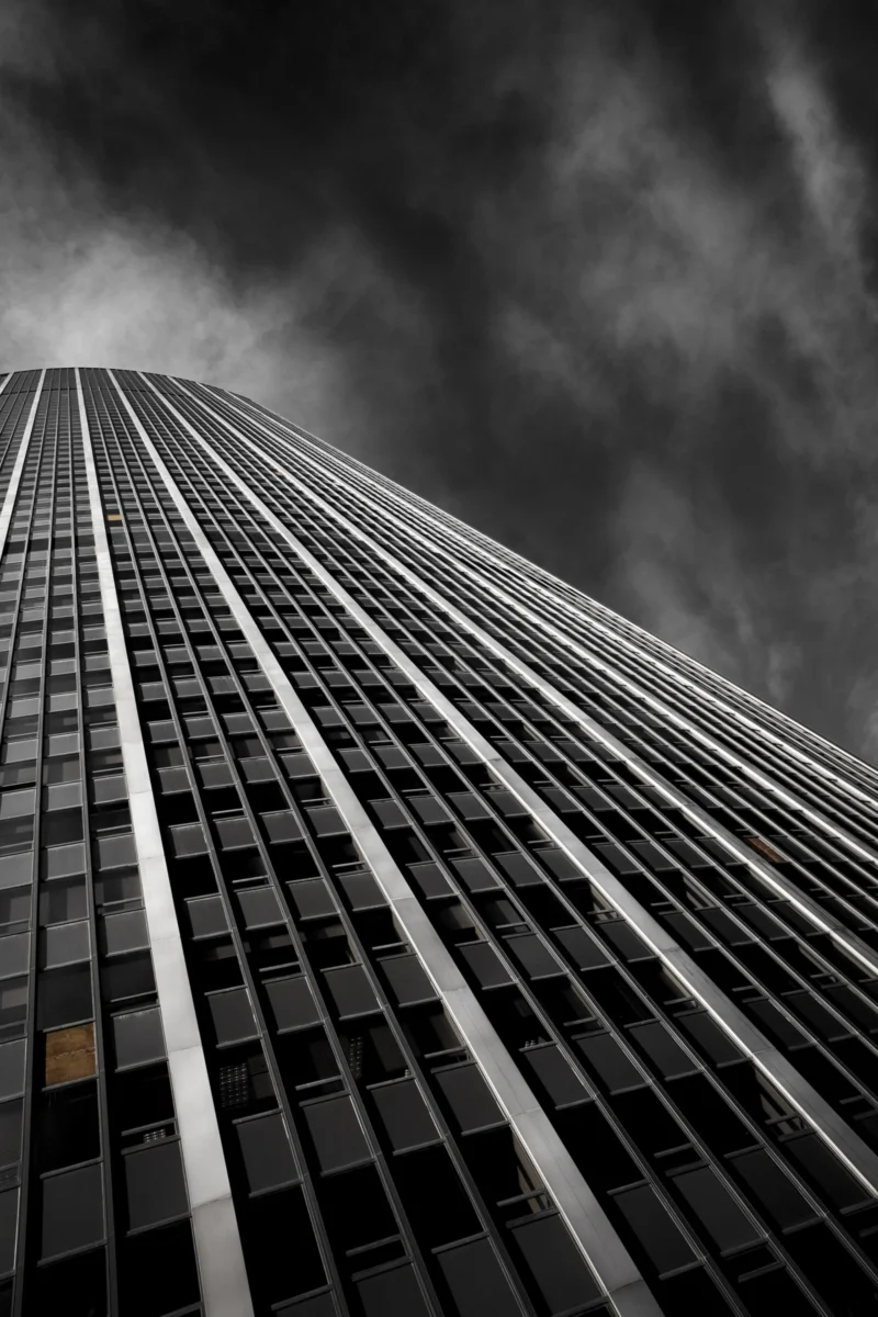 Thierry · 04/07/2025 · 2560 x 3840 Cette photographie met en scène un **gratte-ciel moderne** comme sujet principal. Le **cadrage serré** accentue sa hauteur, avec une perspective ascendante renforçant son impression de verticalité.La **composition** est épurée, centrée sur les lignes architecturales et les jeux d’ombres. La **lumière**, contrastée, crée des effets dramatiques, avec des zones sombres et des reflets sur les vitres.Les **couleurs dominantes** sont les **tons de gris**, renforçant une ambiance **sombre et mystérieuse**. Le style, **minimaliste et urbain**, évoque une atmosphère **industrielle et puissante**, presque menaçante.