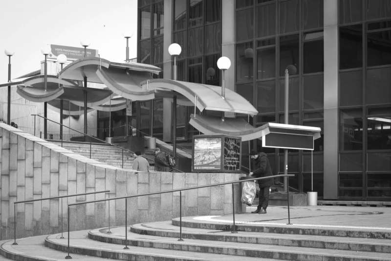 Thierry · 04/07/2025 · 3840 x 2560 Cette photographie en noir et blanc montre un bâtiment moderne avec une structure architecturale distinctive en surplomb. Le sujet principal semble être l'entrée d'un espace public ou culturel, avec des escaliers et une rampe d'accès.La composition met en valeur les lignes géométriques et les jeux d'ombres. Le cadrage inclut des éléments urbains comme des lampadaires et des bâtiments adjacents. La lumière est douce, créant des contrastes marqués entre les zones éclairées et ombragées. L'absence de couleurs accentue les textures et les formes architecturales, renforçant une ambiance à la fois futuriste et intemporelle.