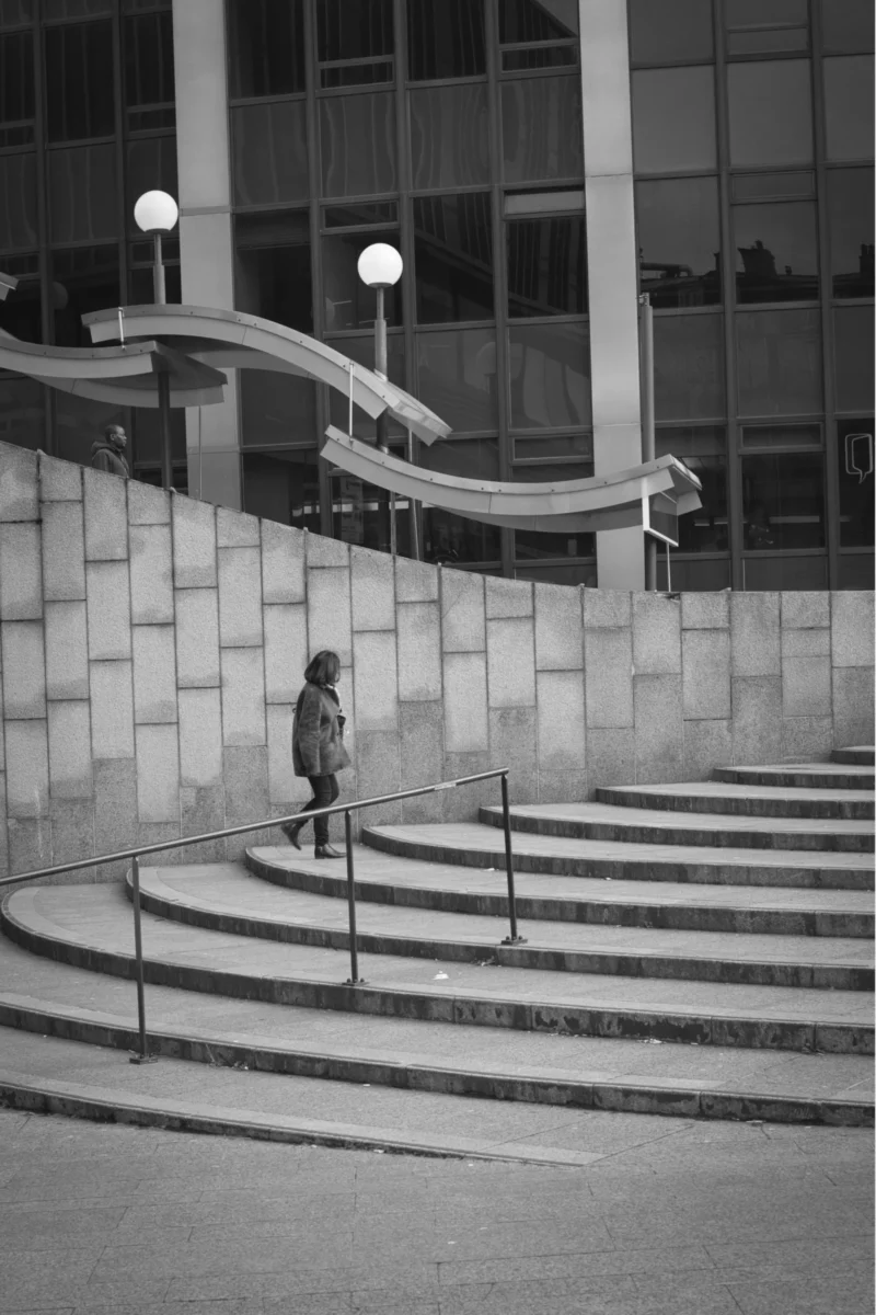 Thierry · 04/07/2025 · 2560 x 3840 Cette photographie en noir et blanc met en scène une personne comme sujet principal, debout sur un escalier extérieur moderne. La composition repose sur des lignes architecturales nettes, avec des marches en demi-cercles et des balustrades. Le cadrage est serré, centré sur la personne et les escaliers, excluant les distractions.La lumière, douce et contrastée, accentue les ombres et les formes géométriques. Les couleurs dominantes sont les nuances de gris, typiques du noir et blanc, renforçant l’aspect minimaliste et contemporain. Le style est épuré, presque cinématographique, créant une ambiance calme et introspective.
