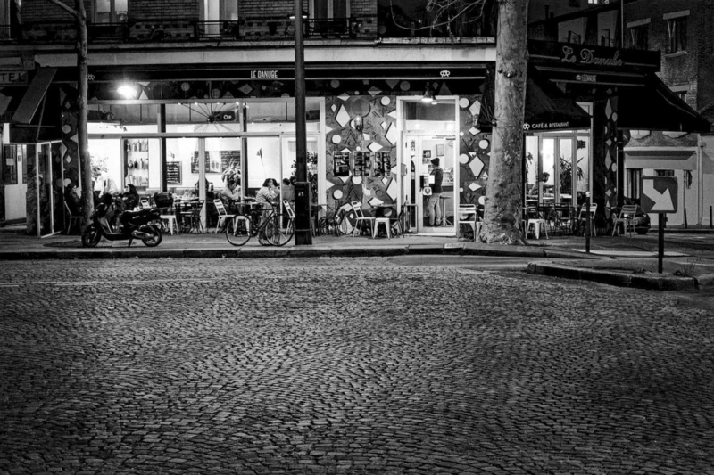 Thierry · 16/01/2024 · 3840 x 2560 Cette photographie en noir et blanc capture une scène nocturne urbaine. Le sujet principal est un café ou restaurant éclairé, avec des clients assis à l'extérieur. La composition met en valeur une place pavée vide en premier plan, encadrée par des arbres et des bâtiments.Le cadrage est légèrement bas, offrant une perspective intéressante. La lumière douce et tamisée crée une ambiance chaleureuse et intimiste. Les contrastes entre ombres et lumières accentuent les détails. Le style est documentaire et poétique, évoquant une atmosphère paisible et légèrement mélancolique. Les tons dominants sont les gris et noirs, renforçant l'effet dramatique.