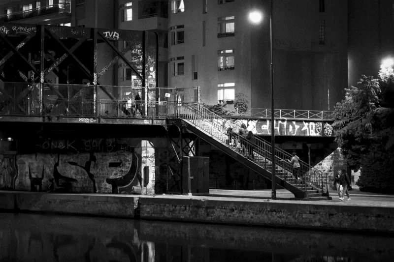 Thierry · 21/03/2024 · 3840 x 2560 Cette photographie en noir et blanc capture un pont urbain de nuit. Le sujet principal est un escalier métallique menant à une passerelle surplombant une voie d’eau, avec des graffitis visibles sur les murs adjacents. La composition met en valeur les lignes architecturales et les jeux d’ombres.Le cadrage inclut des éléments urbains comme des bâtiments éclairés et des structures industrielles. La lumière, tamisée et artificielle, crée des contrastes marqués. Les couleurs dominantes, bien que monochromes, jouent sur les nuances de gris et de noir, renforçant une ambiance mystérieuse et nocturne, typique d’un style photographique documentaire ou urbain.