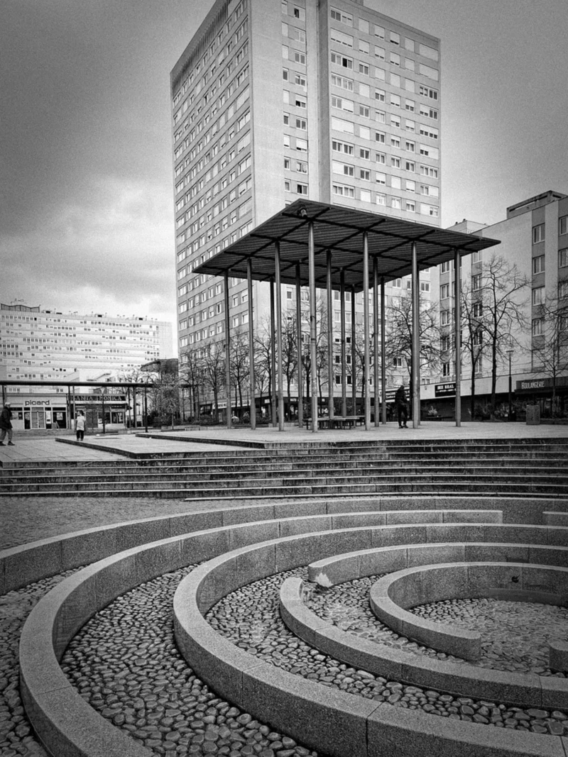Thierry · 24/03/2024 · 2880 x 3840 Cette photographie en noir et blanc met en scène un bâtiment moderne suspendu, sujet principal, soutenu par des piliers. La composition joue sur les lignes courbes des allées pavées en spirale au premier plan, menant vers le bâtiment. Le cadrage est large, incluant une partie du ciel et des arbres dénudés.La lumière, douce et diffuse, crée des contrastes nets entre ombres et lumières. Les teintes dominantes, en niveaux de gris, renforcent la modernité et la géométrie de la scène. Le style architectural minimaliste et épuré confère une ambiance à la fois futuriste et contemplative.